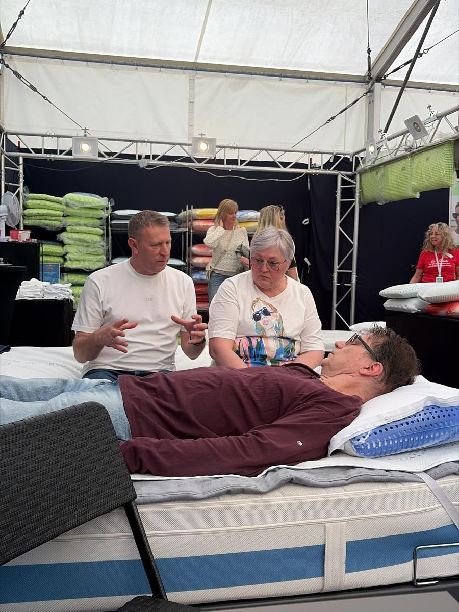 Beratungsgespräch am Bett: ein Mitarbeiter erklärt einem Ehepaar die passende Kissenhöhe