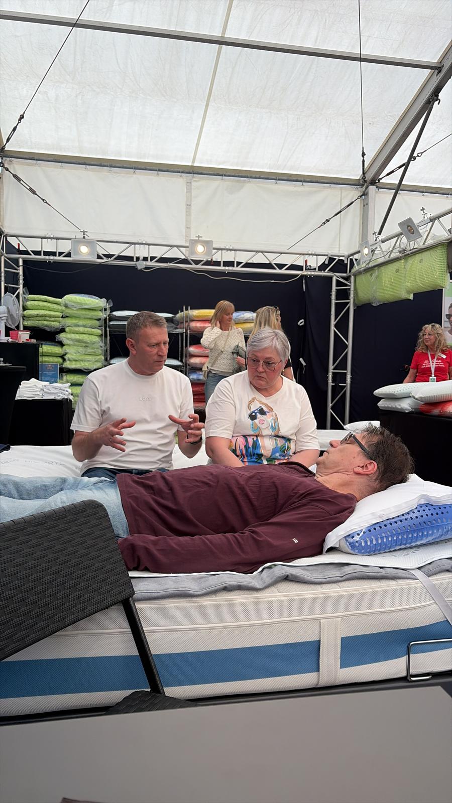 Kunde liegt zum Probeliegen auf dem Bett, waehrend das Schlafzauber-Team die Kissenhoehe erklaert